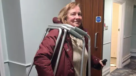 Help Ukraine BAMK UK Community Interest Company (CIC) A woman, holding two silver metal walkers, over her shoulder, smiling at the camera, with long fair hair, wearing a red coat, and cream beige top. She is standing in a corridor that has doors open. 