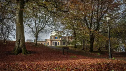 Malcolm MacBain An observatory building is viewed through a gap in autumnal trees which have shed many of their golden brown leaves across the grass in the foreground