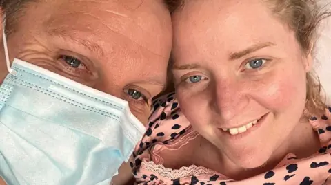 Melinda Tidy and her husband smiling with their heads leaning together. He is wearing a medical mask. She has long blonde hair piled in a bun on her head and blue eyes. He has dark hair and blue eyes. 