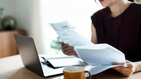 Getty Images A woman sits at her kitchen table looking at paper forms