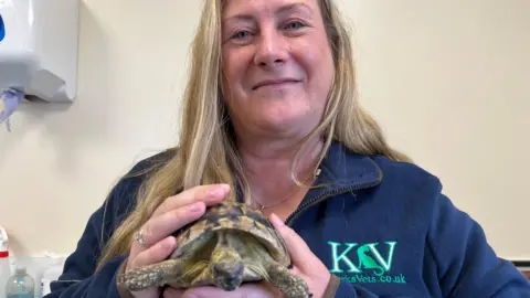 Sara Marchant has long blonde hair with streaks of brown tones. She's wearing a blue fleece and is clutching a tortoise with both hands