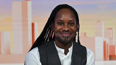 Head shot showing Professor Jason Arday sitting in the Sunday with Laura Kuennsberg studio. He is smiling at the camera with long black locs, wearing a white shirt with a grey waist coat.