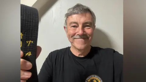 Family handout A man with grey hair and a grey moustache is wearing a black t-shirt with the logo of a taekwondo club on the breast. He is holding a black belt in his hand and smiling. 