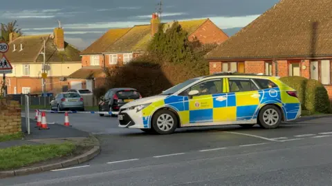 John Nurden A residential road has been cordoned off. There is a police car parked by the road.