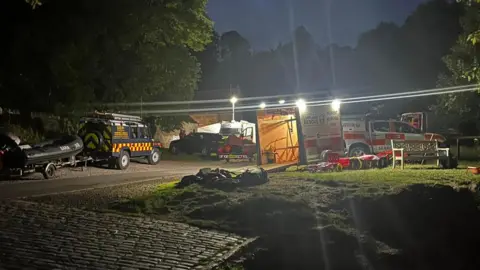 Lifeboats and emergency service vehicles are parked on the edge of a river bank. Floodlights are being used to assist the search.