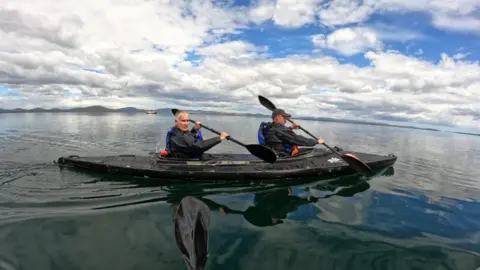 Cockleshell 22 Image shows two men in a kayak on glass like water. They are paddling under a cloud filled blue sky.