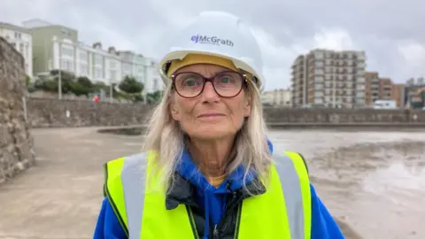 Jan Doyle, a middle-aged woman with grey hair and glasses is wearing a hard hat and high-vis jacket. She is stood next to the marine lake