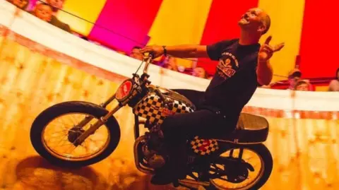 Ken Fox A motorbike rider circles the steep wooden wall of a “wall of death” arena, riding sideways while gripping the handlebars. Spectators lean over the top edge watching as the rider performs close to them under a red-and-yellow striped tent.