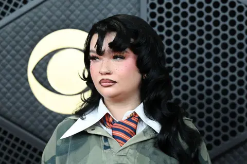 Lola Young has long black hair, she is wearing a dark green jumper with a large white collar over the top, she has a orange and blue striped tie on