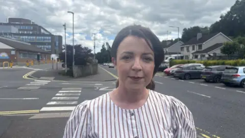 Liz Kimmins, a woman with brown hair, wearing a brown a white striped top. Daisy Hill hospital is in the background, on the left.