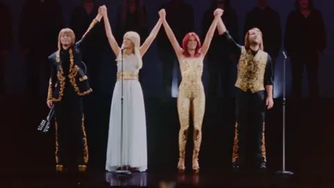 ABBA Voyage The Abba avatars onstage, with the band members clasping their hands together in the air. Bjorn has a black suit with gold trim, Agnetha wears a white flowing dress with a gold belt, Frida wears a gold catsuit, and Benny a black shirt and trousers with gold waistcoat.