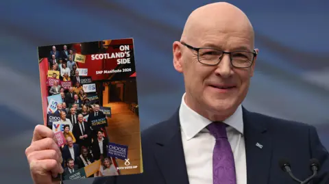 John Swinney, who is bald with glasses, holds a manifesto booklet in his right hand. He is wearing a business suit with a purple tie 