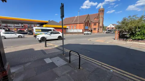 The photo is taken from a street off a main road. Straight ahead is the main road busy with traffic. A petrol station and a church can be seen in distance.