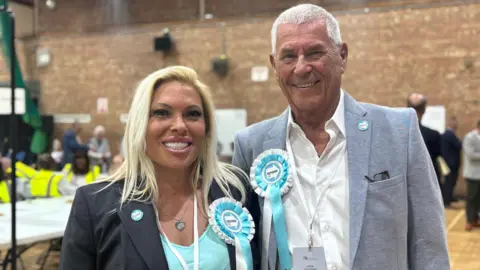 Claire Hamilton/BBC Emma Beck, who has blonde hair, is wearing a turquoise top under a grey blazer, and Tony Keogh, who has white hair, a white shirt and a light grey blazer, smile at the camera in a gymnasium. Both have turquoise and white Reform rosettes pinned to their lapels.