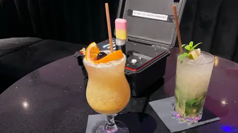 BBC/Gem O'Reilly Two cocktails - one yellow, another looking suspiciously like a mojito, on a table in front of a black box containing a machine which contains a vape