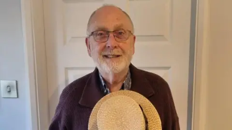 Roger Wash Roger Wash has short grey hair and a beard. He is wearing glasses, a jumper and a shirt while clutching a straw hat to his chest.