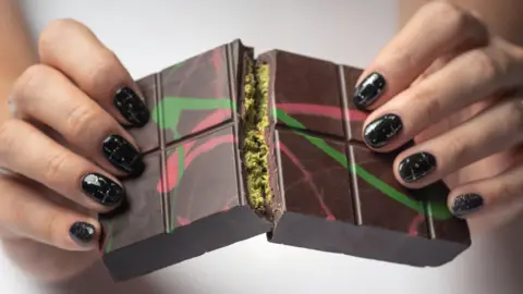 A person with black glittery nails breaks open a bar of Dubai chocolate to show the green filling inside