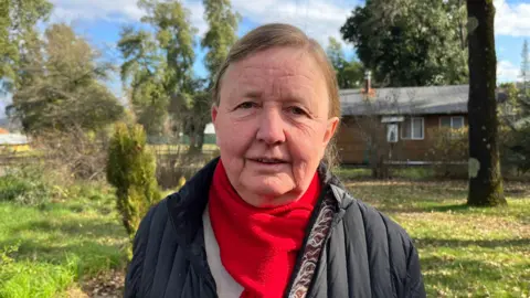 Grace Livingston Eric Timm, dressing in a red scarf, looks at the camera while standing in the villa. Behind it you can see the house and green grass and trees. 