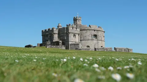 Pendennis Castle stands on a grassy hill dotted with small white flowers, with the stone keep and fortifications set against a clear blue sky.