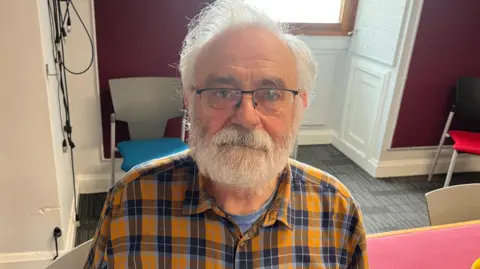 A man with white hair and a white beard, wearing glasses and a yellow checked shirt.