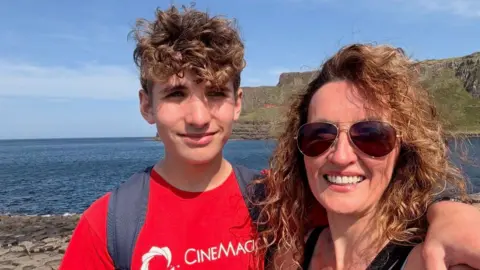 Family Handout A photo of a boy (left) and a woman (right)
The boy is wearing a red tee shirt with the words 'cinemagic' written in white. He has brown curly hair and is smiling at the camera. He is also wearing a black backpack that you can see the straps over his shoulders. The woman is wearing brown sunglasses, with brown curly hair and is smiling at the camera. In the background there is the cliffs and sea. 