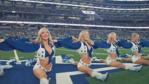 Netflix The Dallas Cowboys Cheerleaders in formation on the football field with crowds behind them. Four blonde cheerleaders look straight ahead with their legs flat, performing the splits.