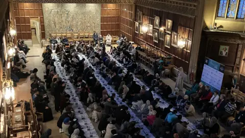 Shariqua Ahmed/BBC People sitting on rows of mats laid on the floor inside the King's College's dining room. It is a large room with a high ceiling and wood panelling.
