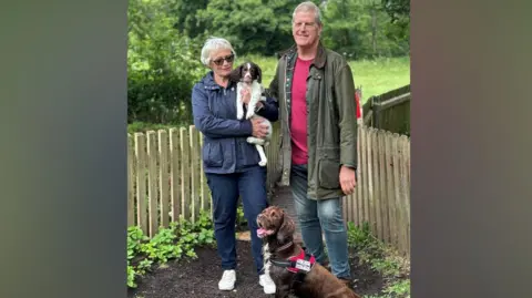 A man and woman with two dogs. The woman, who is stood on the left, is holding a brown and white dog in her arms. A brown dog is stood near them.