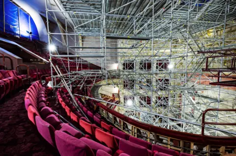 Anneleen Linsday Rows of velvet red seats in the upper circle of a theatre - the stage area is covered with scaffolding towering to the ceiling