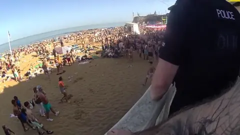 A large mob of young people, mainly men and boys in beach wear, stood on a sunny and busy beach. The picture is taken from a high vantage point with a police bodycam. Another police officer is standing at the railing.