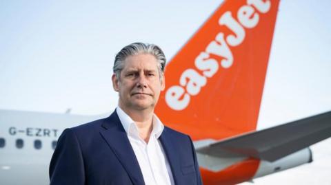 Johan Lundgren with black/grey hair wearing a grey jacket and standing in front of an EasyJet plane