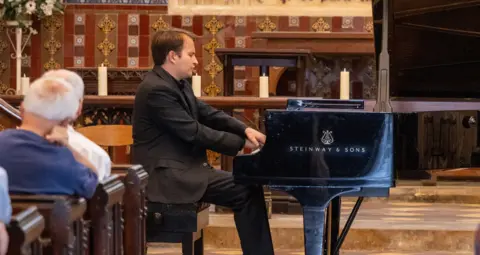 James O'Driscoll Pianist Misha Kaploukhii playing a Steinway piano in Gloucester Cathedral. A number of people are sat in the pews to the side of him watching the performance.