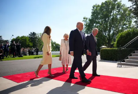 Andrew Harnik / Getty Images Donald Trump and the King walk in front of Melania and Camilla as they walk down the red carpet into the White House, with journalists in the background.