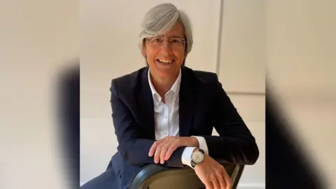 SASH Angela Stevenson looks at the camera and is sitting on a chair and leaning her arms on the back of it. She is wearing a dark suit jacket and a white shirt. She has short grey hair in a  side parting and is smiling at the camera.