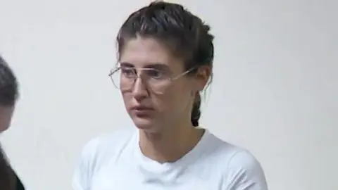 Rustavi 2 Bella Culley filmed in court. She is wearing glasses and has brown hair in a plait and is wearing a white top