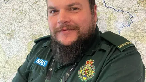 Aaron Wilkes wearing his uniform and lanyard for West Midlands Ambulance Service, which is a green shirt with an NHS badge and an ambulance logo.