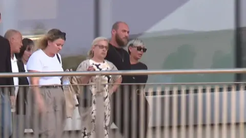 A group of people (men and women) walking past a building. The shot includes five people.