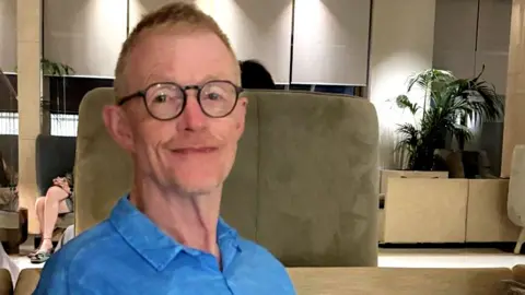Steve Duell Head and shoulders portrait of a man in glasses, smiling gently. He is wearing a blue shirt and is sitting in an area which could be a hotel or office lobby.