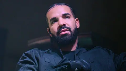 O rapper canadense Drake, da Getty Image, está vestido com camisa, jaqueta e luvas e segura um microfone enquanto olha para a esquerda.