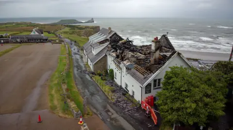 Athena A wide image of the damage the fire has caused to The Worm's Head Hotel. The roof of the hotel has completely collapsed. Behind the hotel is a choppy sea and in front of it is a large empty car park. The picture has been taken on a cloudy day. 