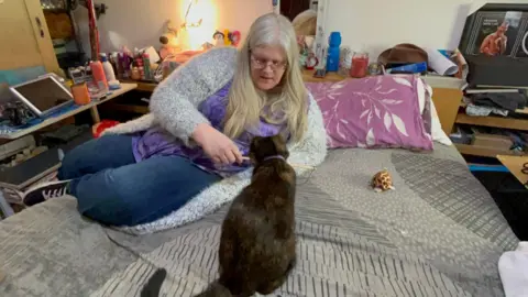 Tanya Clifton on her bed in her bedroom, playing with a black and brown cat. There are various personal items on the shelves around the bed. She is looking down as she plays with the cat.