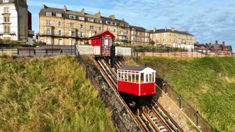 Alistair A red and white cliff lift nearly at the top of the track. There is a red shed at the top which is in front of a road. Behind the road are large, terraced houses made from beige bricks. The bank on either side of the lift is grassy and the sky above is blue with small, grey clouds.