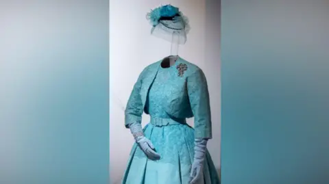 PA Media The turquoise blue dress and Bolero Jacket as they are displayed in the Royal Collection exhibition with matching light blue gloves and diamond bracelet and fascinator.