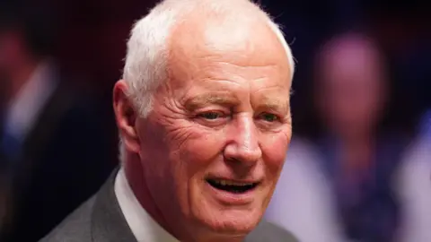 Barry Hearn is wearing a grey suit jacket and white shirt. He has short white hair.