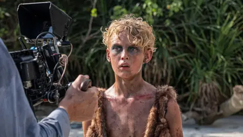 BBC/Eleven/J Redza A blonde haired boy wearing a fur jacket with mud on his face looks towards a camera, he's standing on the beach of an island.
