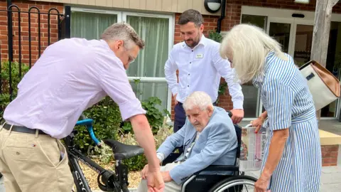 A group of people are looking at a bicycle which is in the forecourt of a care home. One person, who is on the left, is holding and pointing at the bike. The others are around him looking at it.