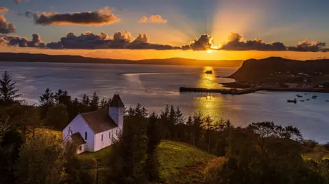 Andrew Briggs Sun setting over a tranquil bay, surrounded by trees with a white building perched on the hillside overlooking the water.