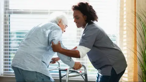 An elderly man in a shirt and jeans is helped up out of a chair by a nurse. The man is holding a walking frame and is positioned by a widow. The nurse has a grey uniform top on and dark curly hair.