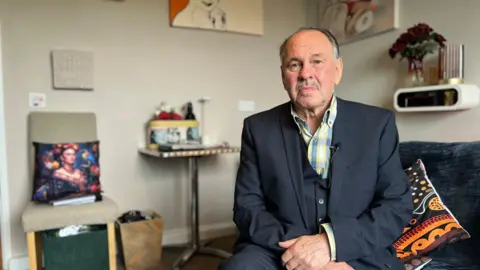 Jeff Thomson from Penrith is sitting on a settee, wearing a grey three-piece suit. There is a chair and table behind him.