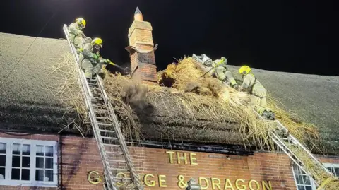 Four firefighters standing at the top of silver ladders, leaning up against the thatched roof of the George and Dragon pub. It is a red brick building with sash windows and a tall chimney in the middle. The sky is dark and the fire is extinguished, but the thatched roof has been torn and burnt.